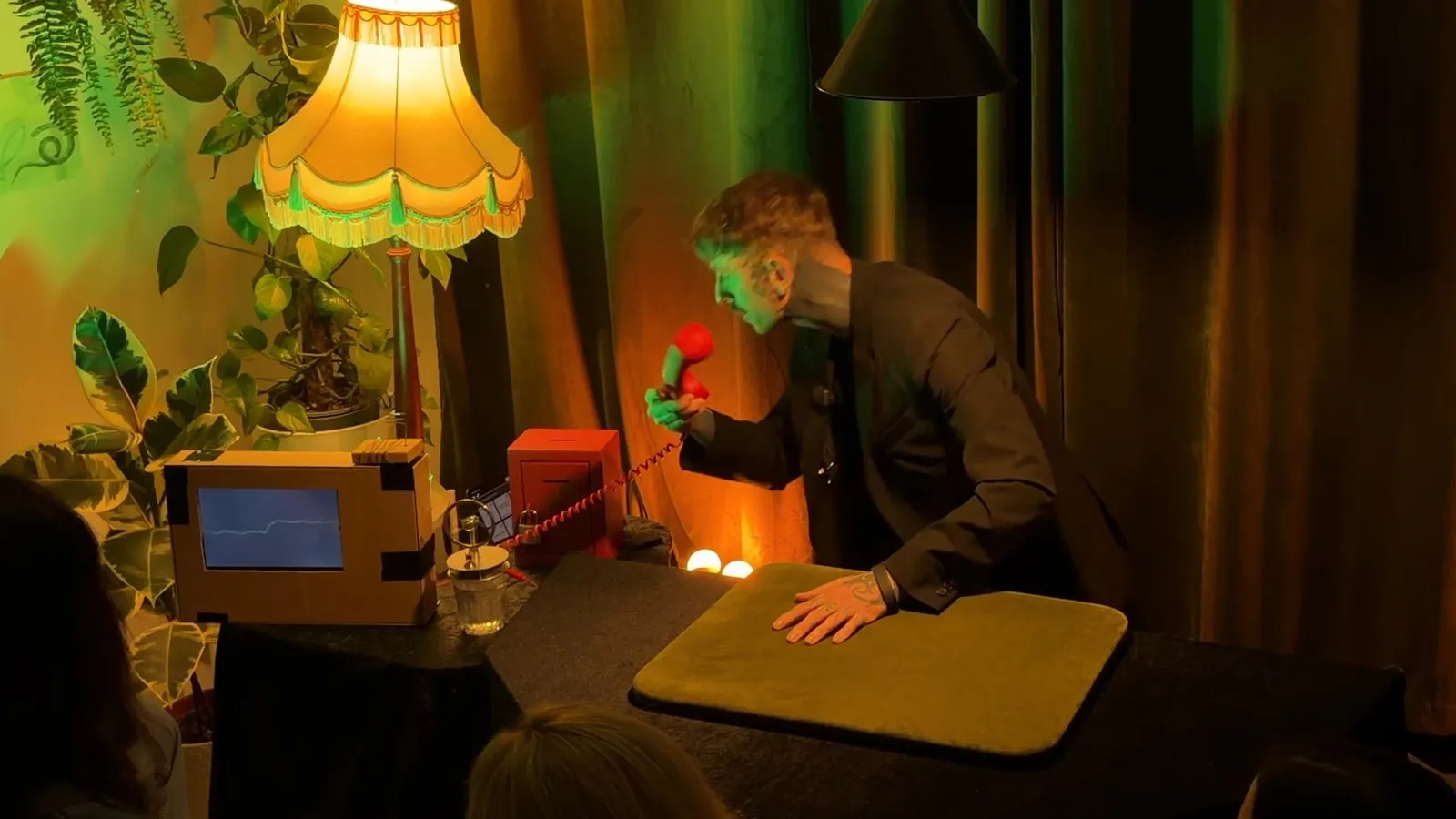 Simon at the table under the yellow lamp, holding up a small red prop